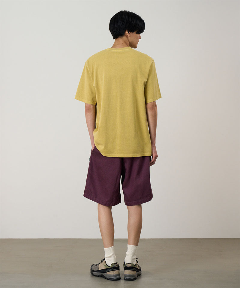 Rear view of a model wearing a sulpher Gramicci G-Scribble Tee Pigment Dyed shirt with purple shorts on a light tone background.
