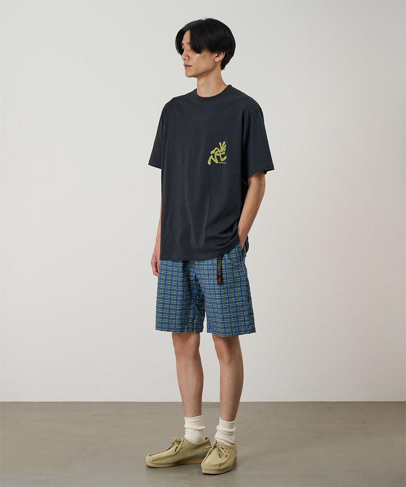Person wearing a black t-shirt with a yellow logo and plaid shorts on a plain background