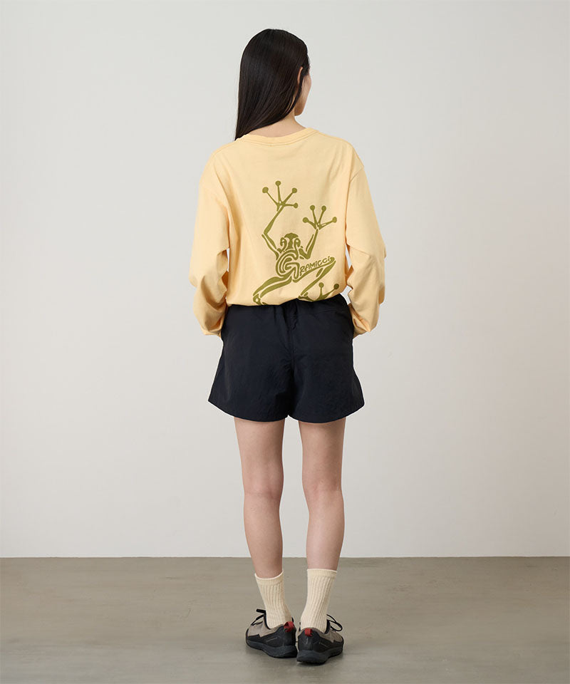 Rear view of a model wearing a mango colored Gramicci Sticky Hands L/S Tee with a green frog graphic and black shorts on a light tone background.