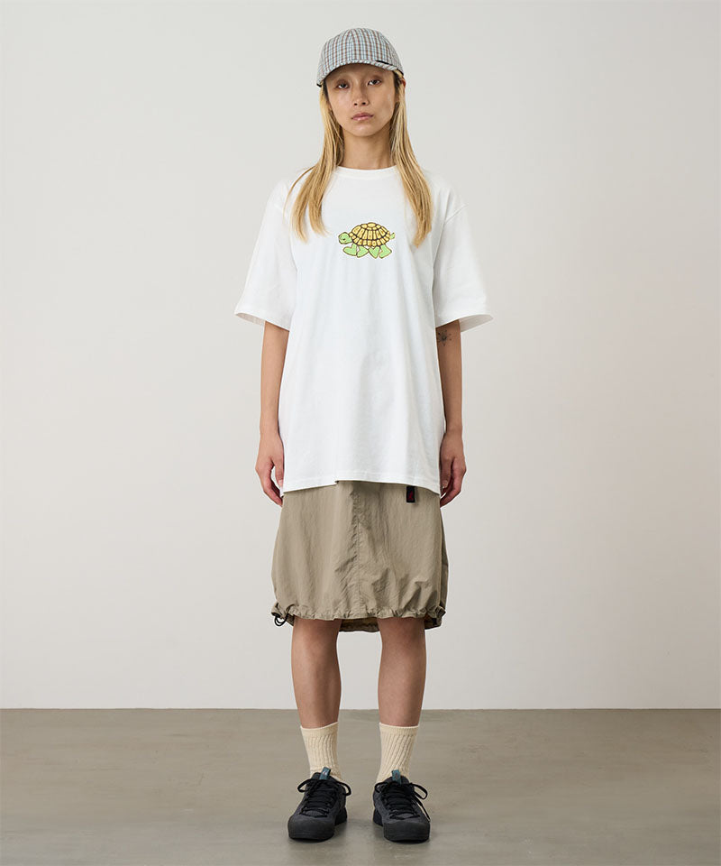Model wearing a white Gramicci Turtle Tee with a a turtle graphic and beige skirt on a light tone background.