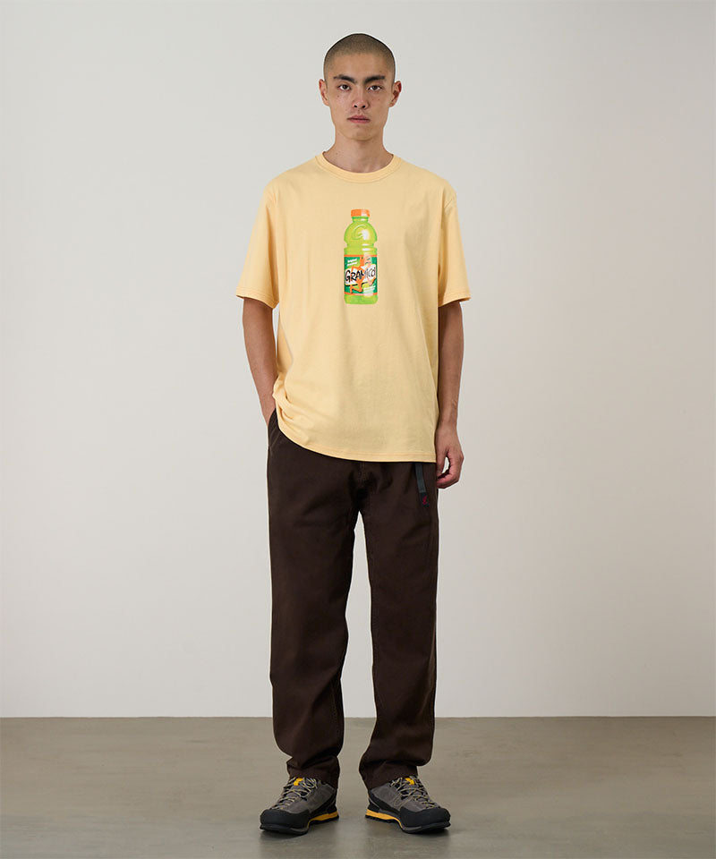 Model wearing a mango Gramicci Quencher Tee with a hydration drink graphic and blue pants on a light tone background.