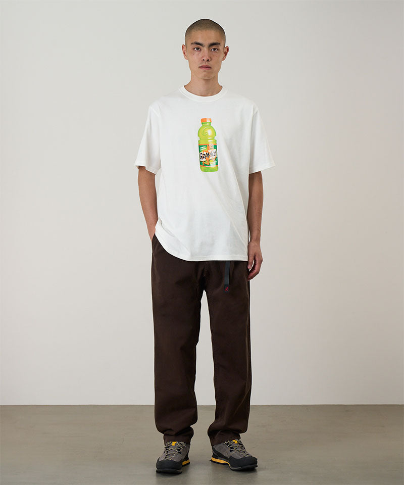 Model wearing a white Gramicci Quencher Tee with a hydration drink graphic and brown pants on a light tone background.