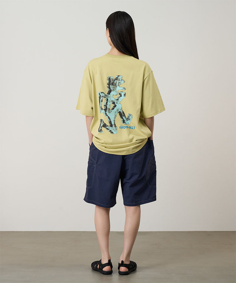 Back view of a model wearing a lime colored Gramicci Ice Sculpture Tee with a a running man graphic and blue shorts on a light tone background.