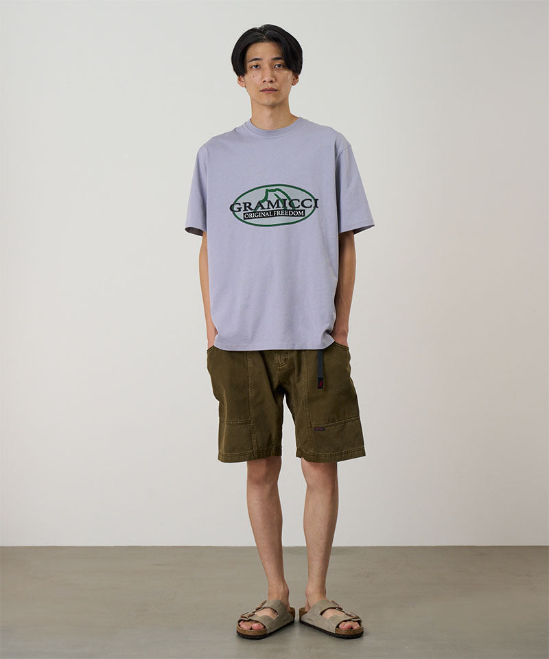 Model wearing a lavender Gramicci Half Dome Tee with a grey and green graphic and green shorts on a light tone background.