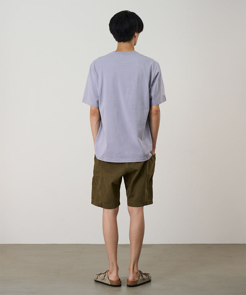 Rear view of a model wearing a lavender Gramicci Half Dome Tee with green shorts on a light tone background.