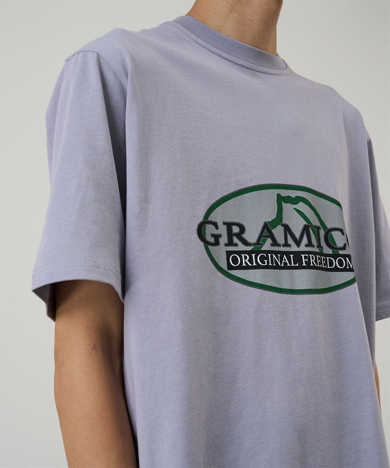 Model wearing a Gramicci lavender Half Dome Tee with grey and green graphic against a light tone background.
