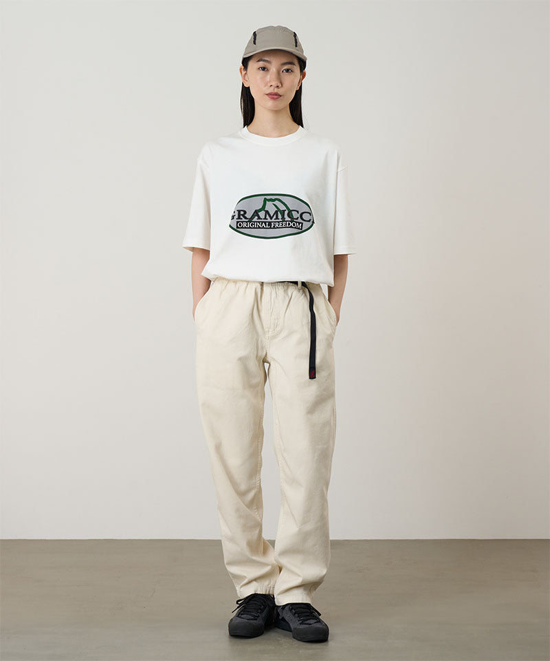Model wearing a white Gramicci Half Dome Tee with a grey and green graphic and light beige pants on a light tone background.