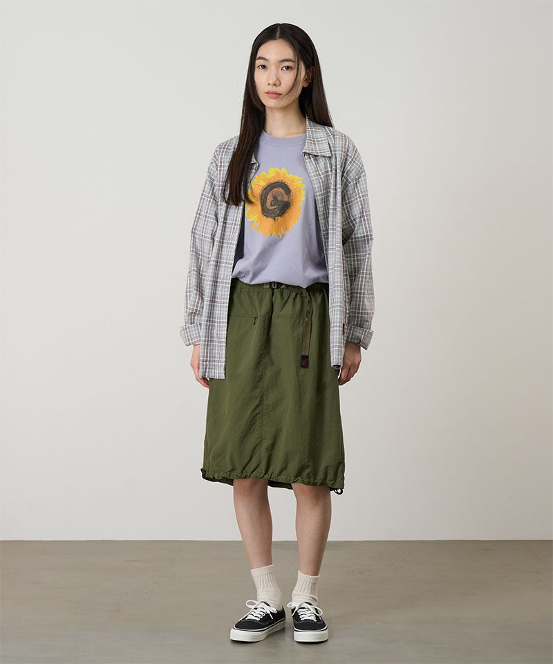 Person wearing a plaid shirt, gray t-shirt with a sunflower design, and green skirt on a plain background