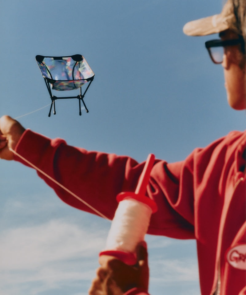 Person flying a mesh camo Gramicci & Helinox Chair One kite against a blue sky.