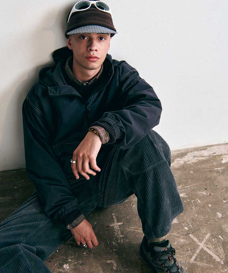 Model wearing a Gramicci black hooded jacket, dark corduroy pants, dark trail shoes, and a fleece brimmed cap with sunglasses.
