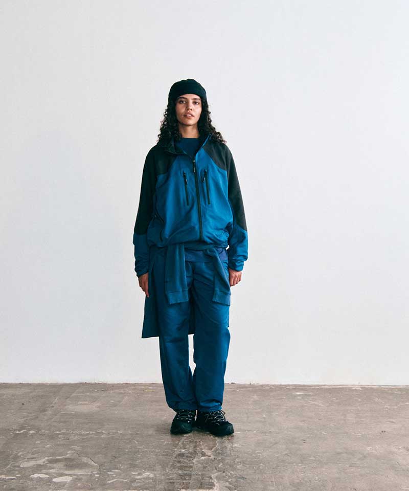 Model wearing a Gramicci blue and black colorblock zip-up jacket, matching teal pants with a sweatshirt tied around the waist, a black beanie, and black trail shoes.