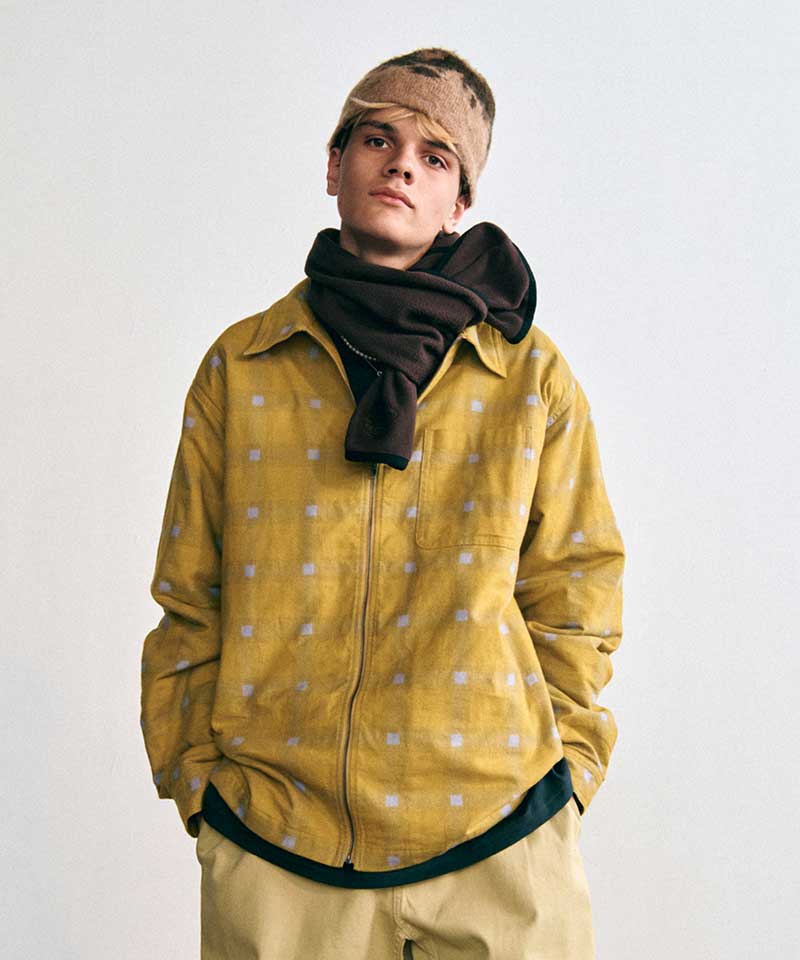 Model wearing a Gramicci yellow geometric-pattern zip-up shirt, a dark brown fleece scarf, a patterned fleece beanie, and tan pants.