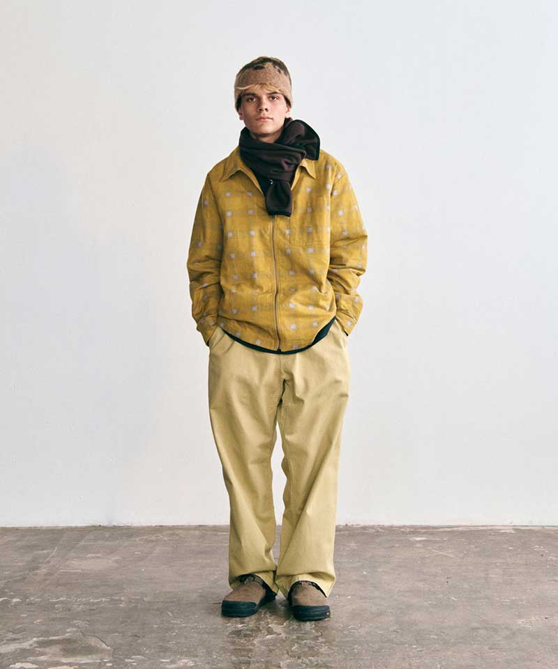 Model wearing a Gramicci yellow geometric-pattern zip-up shirt, a dark brown fleece scarf, a patterned fleece beanie, tan pants, and brown outdoor shoes.