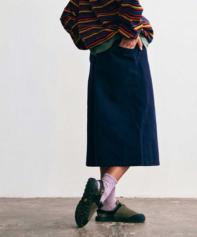 Model wearing a Gramicci navy midi skirt, a multicolored striped sweater, lavender socks, and olive-green outdoor sandals.