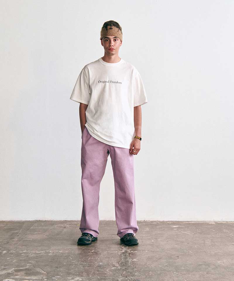 Gramicci white "Original Freedom" graphic T-shirt and relaxed-fit lilac colored pants worn by a person standing against a white studio background.