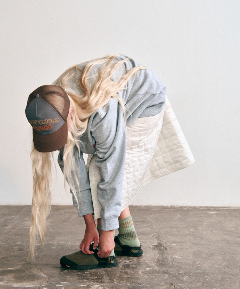 Model wearing a Gramicci brown trucker cap with a "Livin' Under a Rock" graphic, a gray sweatshirt, a cream quilted skirt, green ribbed hemp socks, and olive-green outdoor sandals.
