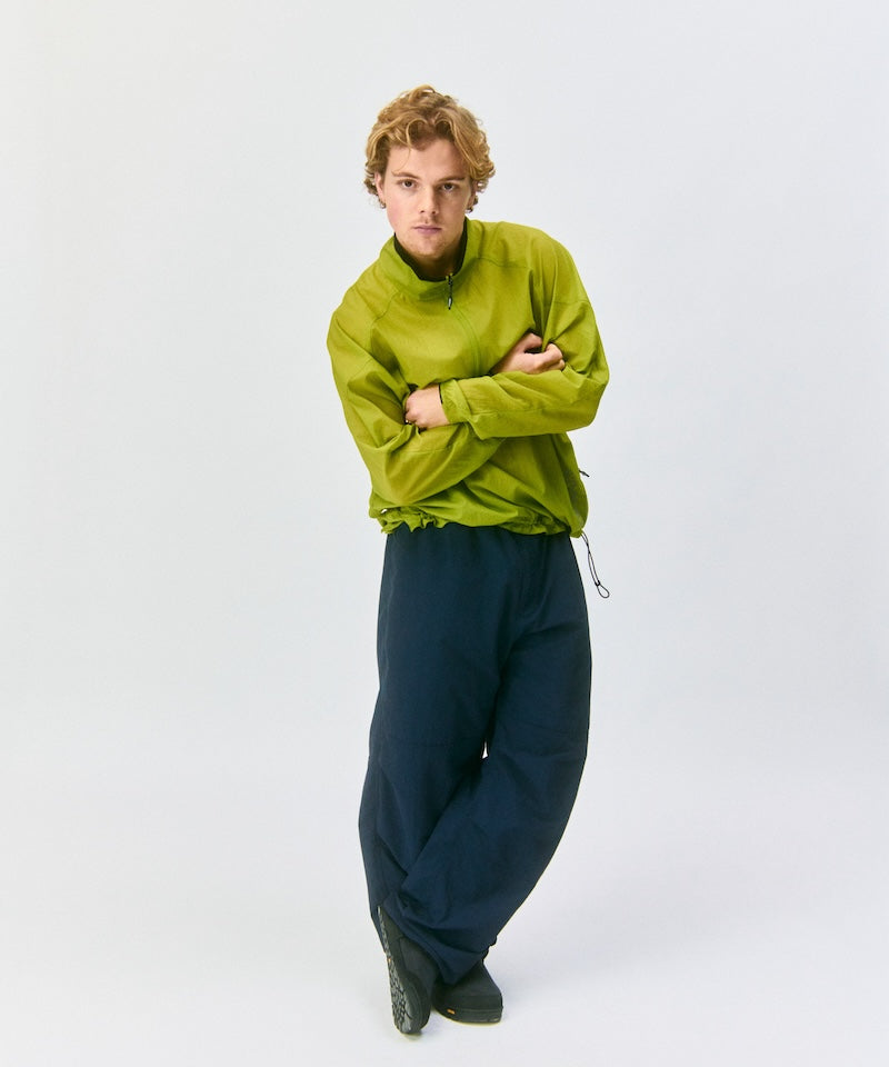 Model wearing a kiwi Gramicci Ultra-Light Packable Jacket and blue pants on a light-tone background.