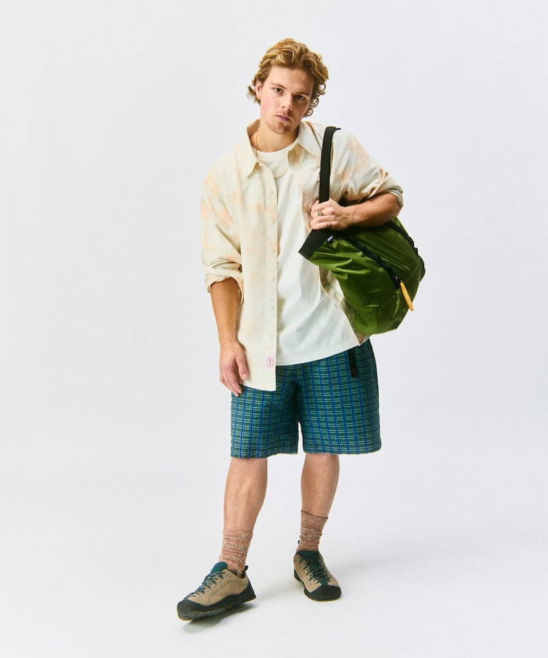 model wearing a beige shirt and blue plaid shorts, holding a green bag on an off-white background.