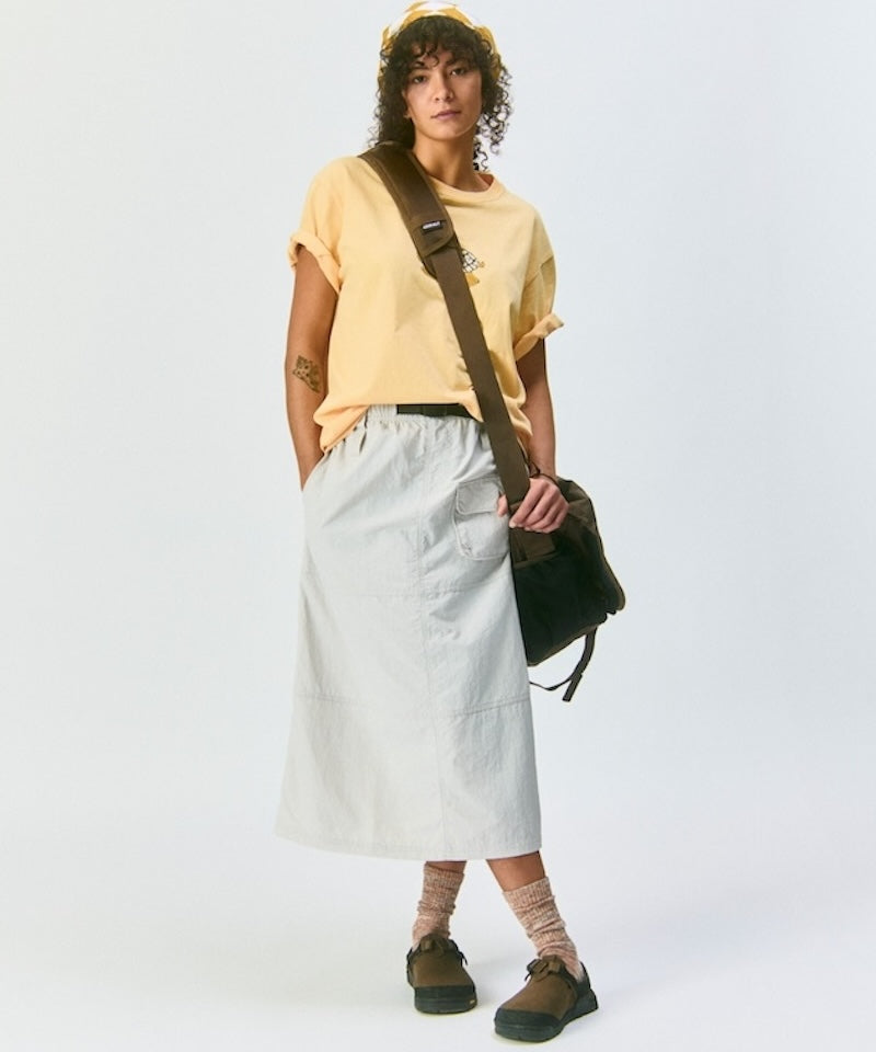 Model wearing a yellow Gramicci t-shirt, light gray skirt, and brown shoes on a light background.
