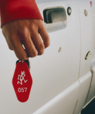 Hand holding a red Gramicci Hotel Keychain with a white Gramicci Running Man logo and number '057' in front of a white vehicle.