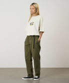 A model wearing olive green pants, a cream short-sleeve top with a Gramicci logo, and black low-top sneakers, standing against a plain light gray studio background.