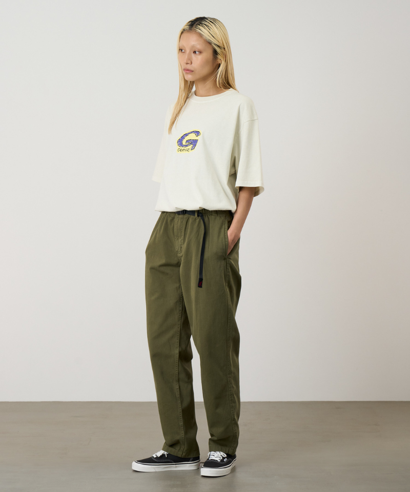 A model wearing olive green pants, a cream short-sleeve top with a Gramicci logo, and black low-top sneakers, standing against a plain light gray studio background.