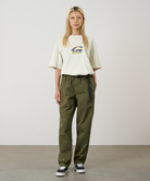 A model wearing olive green pants, a cream short-sleeve top with a Gramicci logo, and black low-top sneakers, standing against a plain light gray studio background.