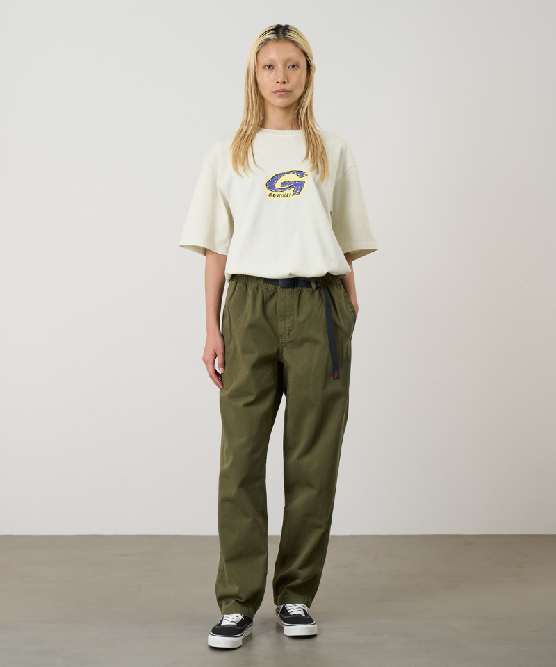 A model wearing olive green pants, a cream short-sleeve top with a Gramicci logo, and black low-top sneakers, standing against a plain light gray studio background.