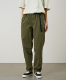 A person wearing olive green pants, a cream short-sleeve top, and black low-top sneakers, standing against a plain light gray studio background.