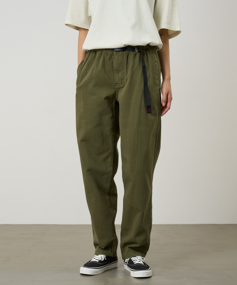A person wearing olive green pants, a cream short-sleeve top, and black low-top sneakers, standing against a plain light gray studio background.
