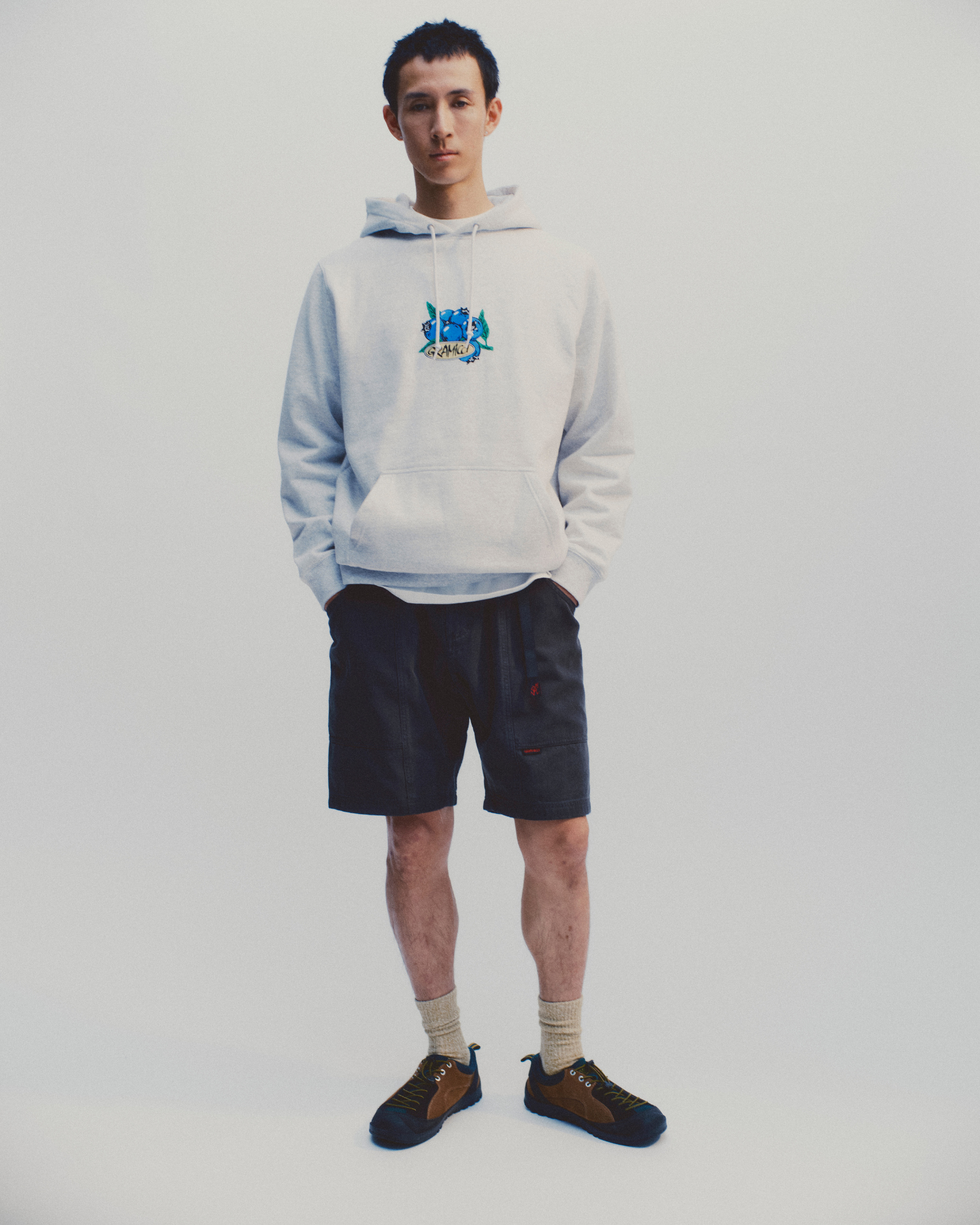 Model wearing a heather ash Gramicci Blueberry Hooded Sweatshirt and navy shorts on a light-tone background.