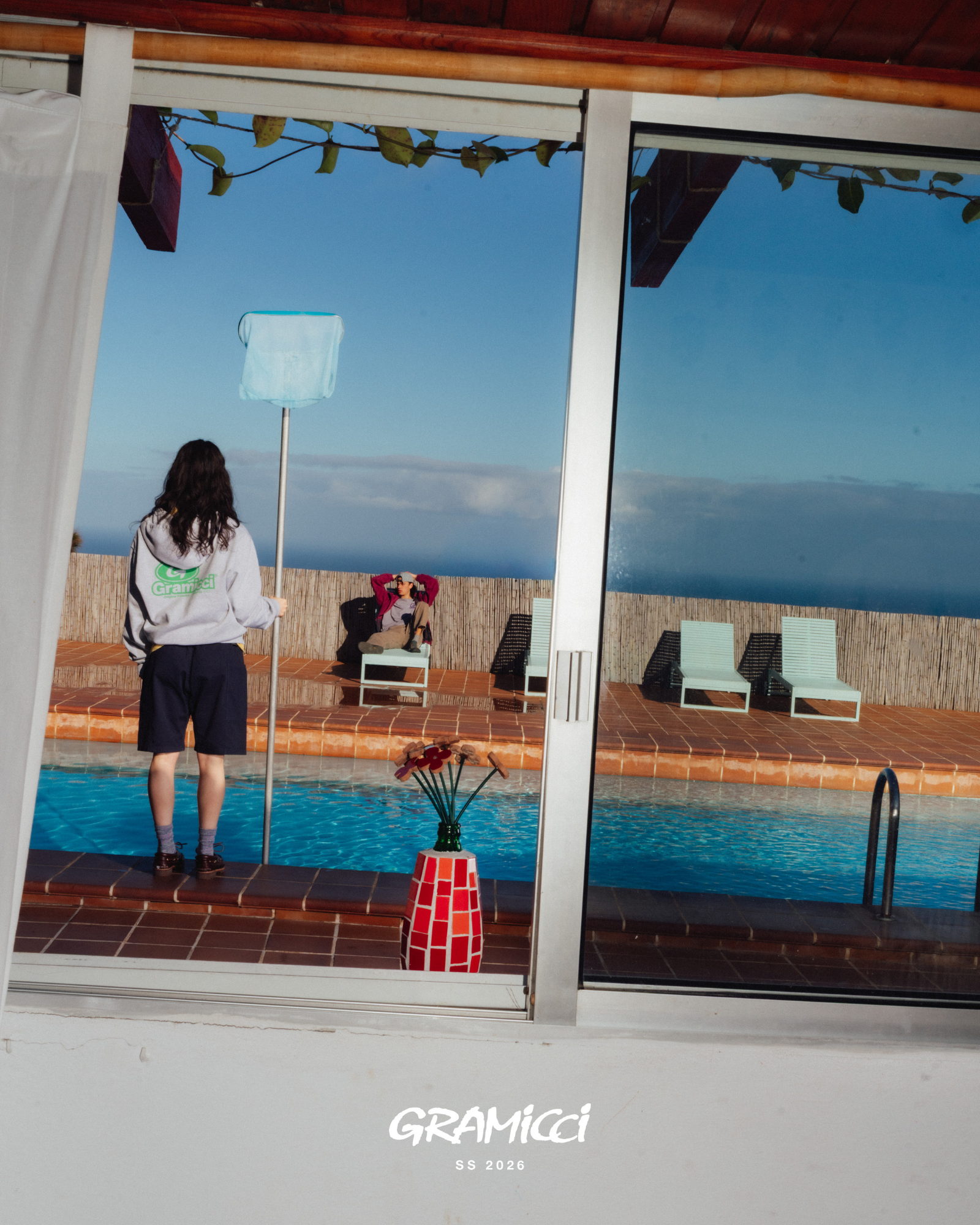 Person standing by a pool with chairs and a clear sky, reflected in a glass door.
