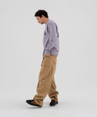 Model wearing beige Gramicci Voyager Pants and a lavender Gramicci Equipped Dirt Long-Sleeve-Tee on a light-tone background.