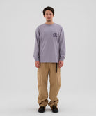 Model wearing beige Gramicci Voyager Pants and a lavender Gramicci Equipped Dirt Long-Sleeve-Tee on a light-tone background.