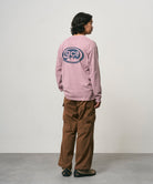 Back view of a model wearing a lilac pigment Gramicci GCE Long Sleeve Tee Pigment Dyed with a graphic and brown pants on a light-tone background.