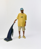 Model wearing blue plaid Gramicci Long Bill Cap and a yellow shirt and green shorts pushing a vaccuum on a light-tone background.