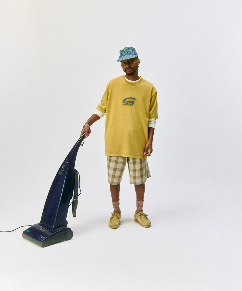 Model wearing blue plaid Gramicci Long Bill Cap and a yellow shirt and green shorts pushing a vaccuum on a light-tone background.