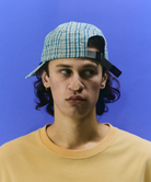 Model wearing olive Gramicci Millwood Cap and a yellow shirt on a blue background.