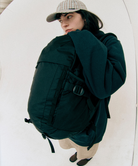 Model wearing green Gamicci Day Pack 25L and a green hoodie on a light-tone background.