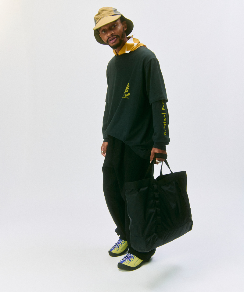 Model carying an olive Gamicci Trail Tote, wearing a green shirt and black pants on a light-tone background.