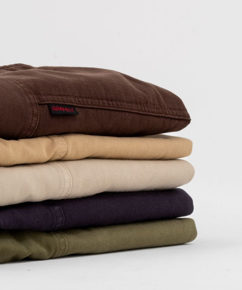 Several folded Gramicci Gadget Pants in various colors with a black belt on a white background
