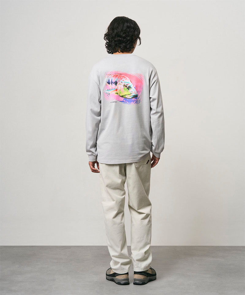 Back view of a model wearing a mist colored Gramicci Bass Long-Sleeve Tee with a graphic and off-white pants on a light-tone background.