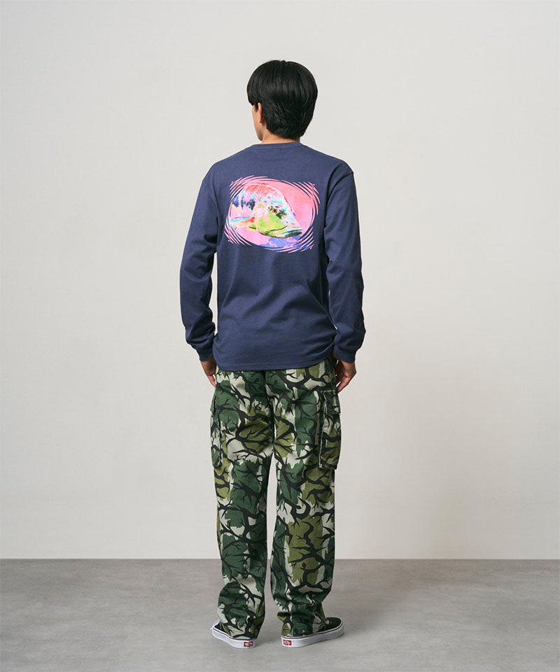 Back view of a model wearing a navy Gramicci Bass Long-Sleeve Tee with a graphic and camo pants on a light-tone background.