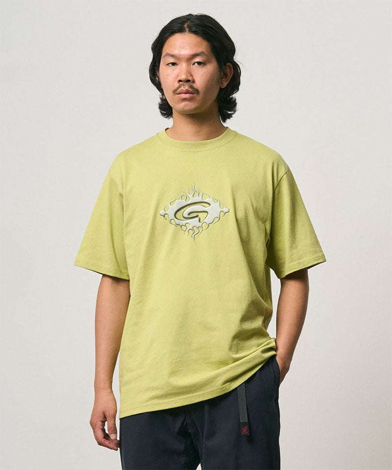 Model wearing a sweet pea colored Gramicci Flames Tee with a graphic and black pants on a light-tone background.