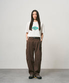 Model wearing a white Gramicci Flames Tee with a graphic and brown pants on a light-tone background.