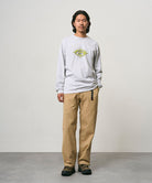 Model wearing a ash heather colored Gramicci Flames Long Sleeve Tee with a graphic and beige pants on a light-tone background.