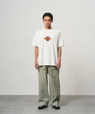 Model wearing a white Gramicci Ladybug Tee with colorful graphic and light green pants on a light-tone background.