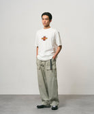Model wearing a white Gramicci Ladybug Tee with colorful graphic and light green pants on a light-tone background.