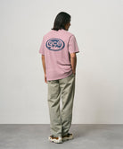 Back view of a model wearing a lilac pigment colored Gramicci GCE Tee Pigment Dyed with a graphic and olive green pants on a light-tone background.