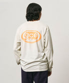 Back view of a model wearing a natural pigment colored Gramicci GCE Tee Pigment Dyed with a graphic and black pants on a light-tone background.